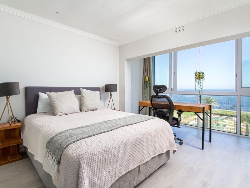A bright, modern bedroom featuring crown molding, recessed lighting, and expansive windows offering panoramic ocean views.