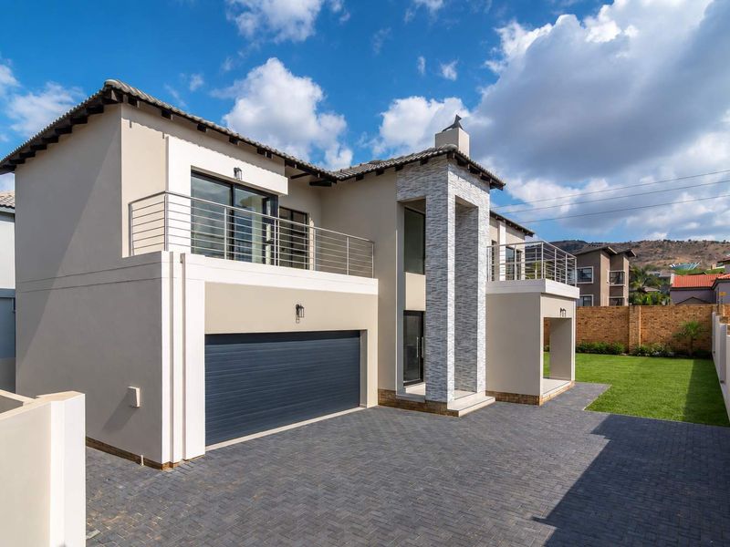 Modern double-story residence featuring a paved driveway, double garage, stone-clad entrance pillar, and upper-level balconies with stainless steel railings.