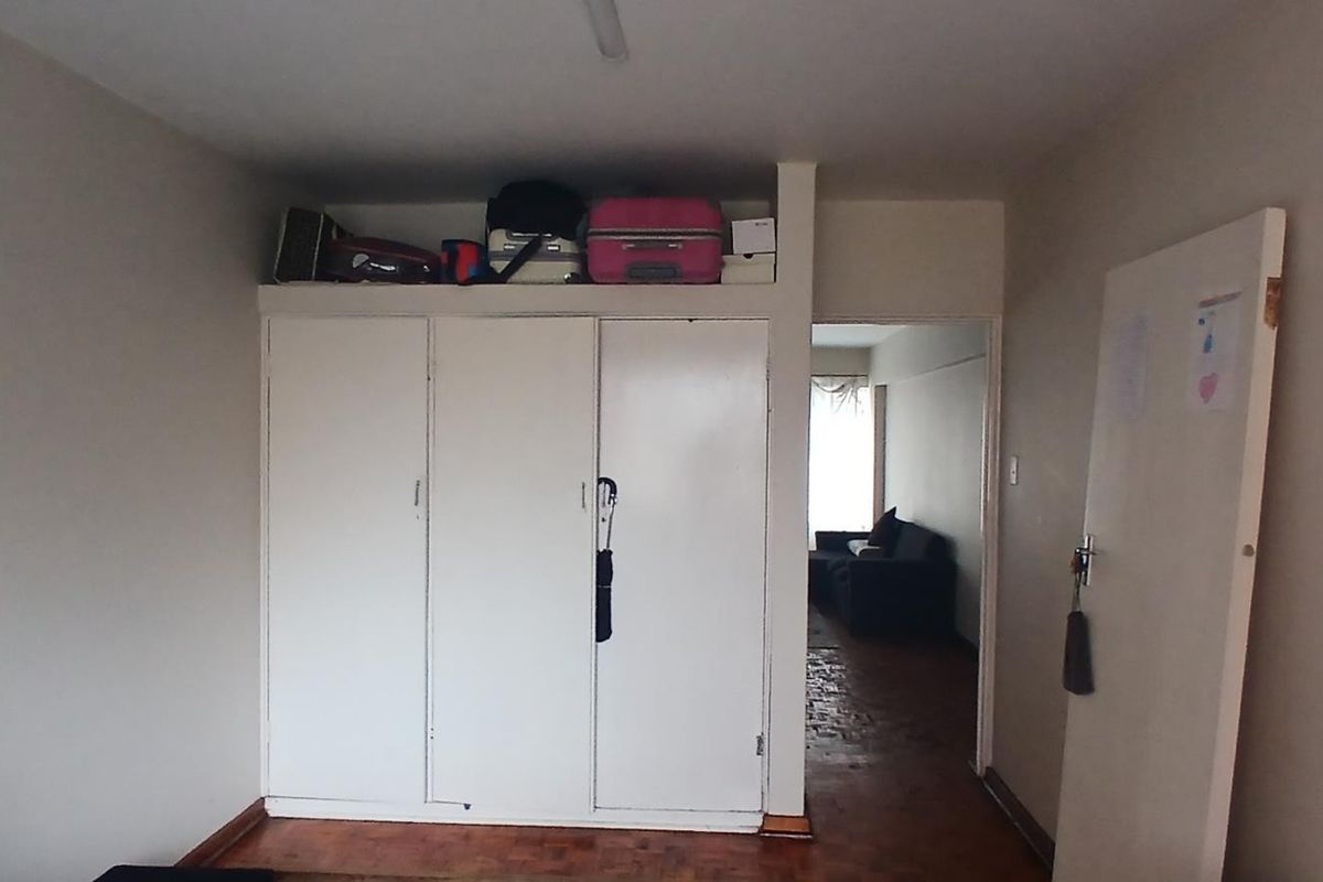 A bedroom featuring a built-in wardrobe with overhead storage and parquet flooring, providing a functional space with direct access to the living area.