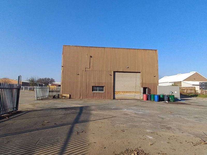 Industrial warehouse facility featuring a large roller shutter door, corrugated metal cladding, and a spacious paved yard area enclosed by security fencing.