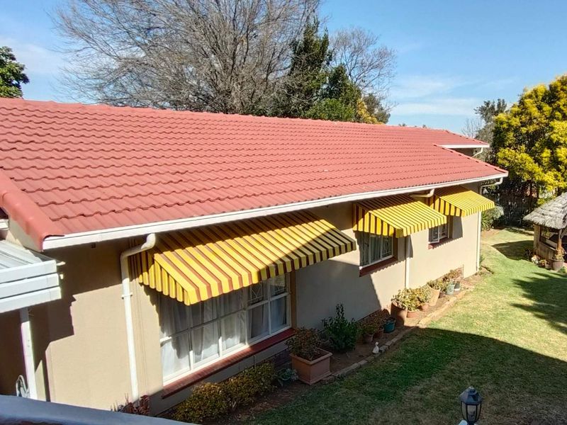 Rear view of the property featuring a tiled roof, striped window awnings, and a detached thatched lapa set within a landscaped garden.