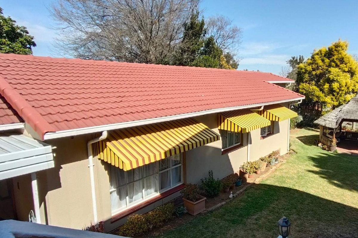 Rear view of the property featuring a tiled roof, striped window awnings, and a detached thatched lapa set within a landscaped garden.