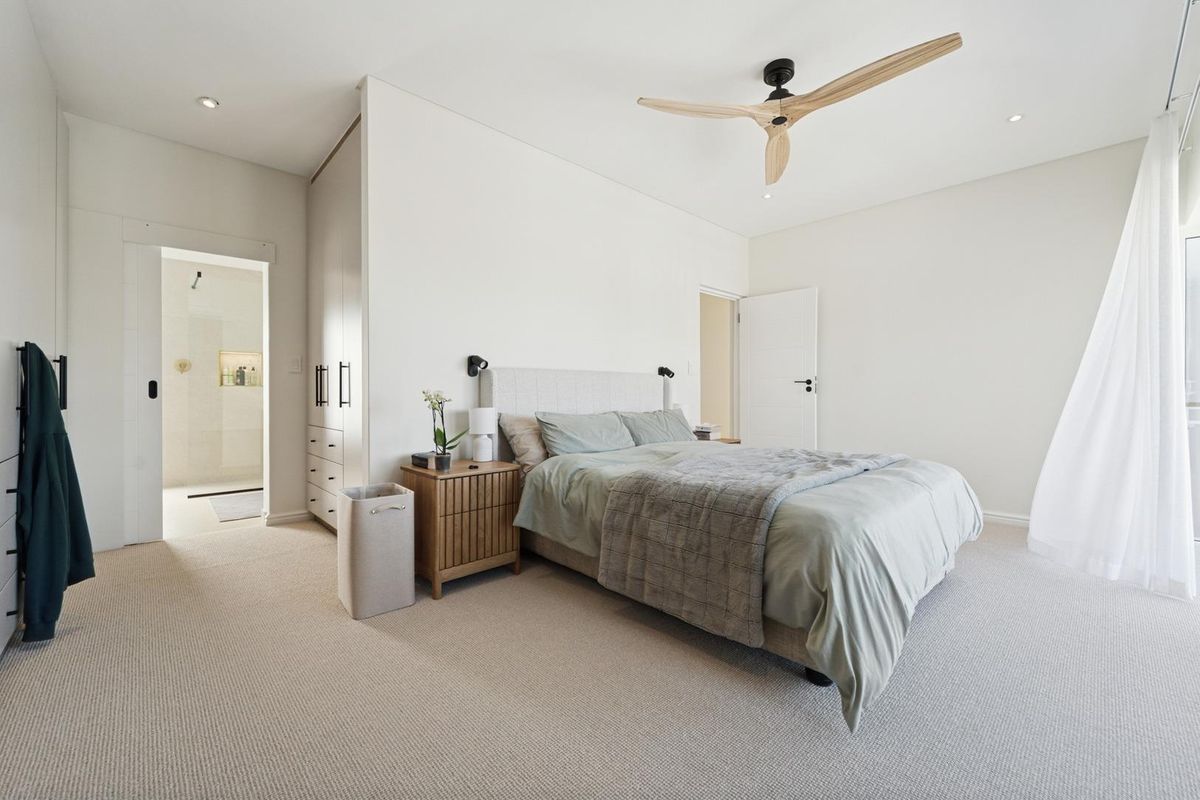 A bright and modern bedroom featuring neutral carpeting, built-in cabinetry, a contemporary ceiling fan, and an en-suite bathroom.