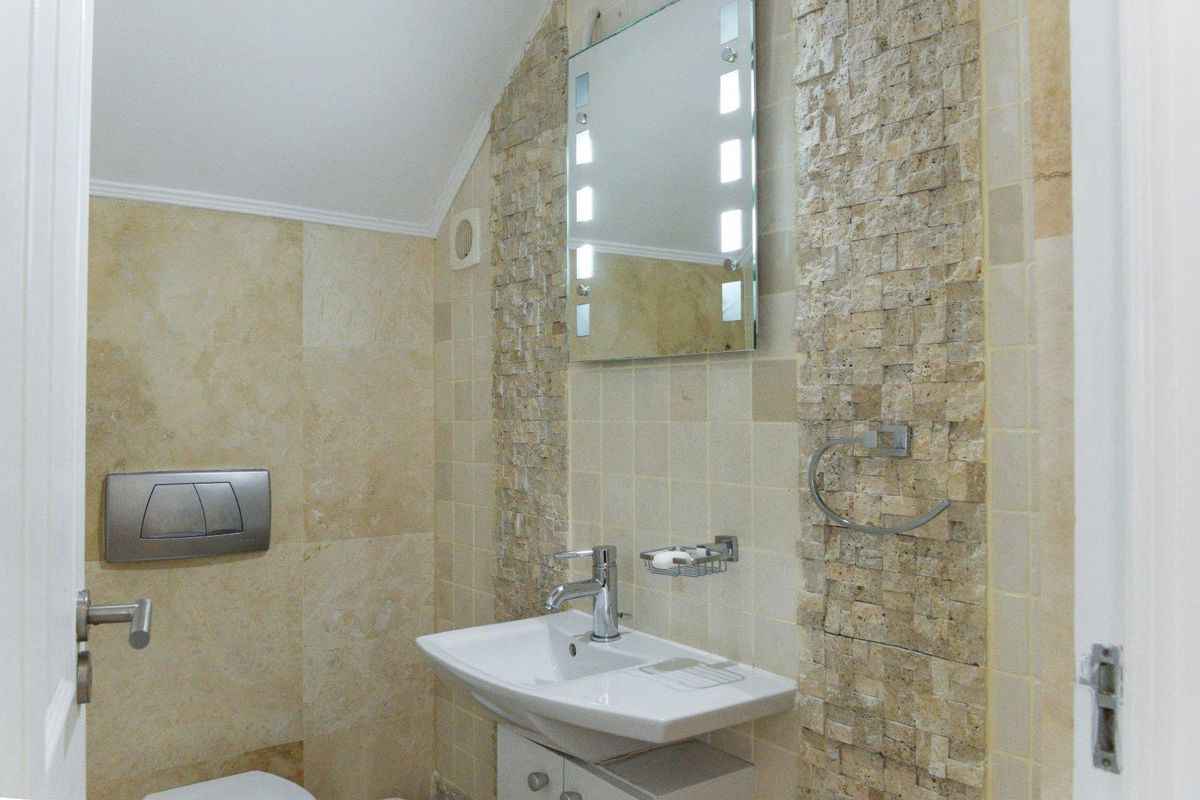 Modern bathroom featuring stone-clad feature wall, wall-mounted toilet, vanity basin, and illuminated mirror with neutral tiling.