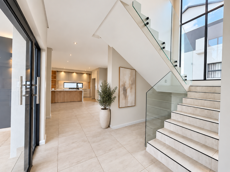 Modern entrance hall featuring a sleek glass balustrade staircase, large format floor tiles, and an open-plan view leading into a contemporary kitchen area.