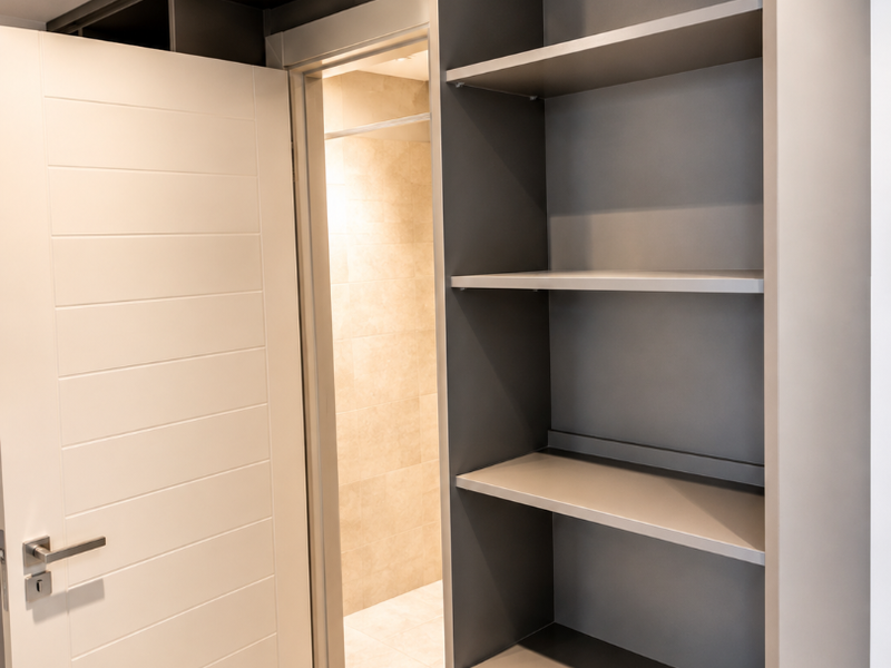 A modern bedroom featuring built-in grey shelving, recessed lighting, and a clean, contemporary door leading to an adjacent tiled space.