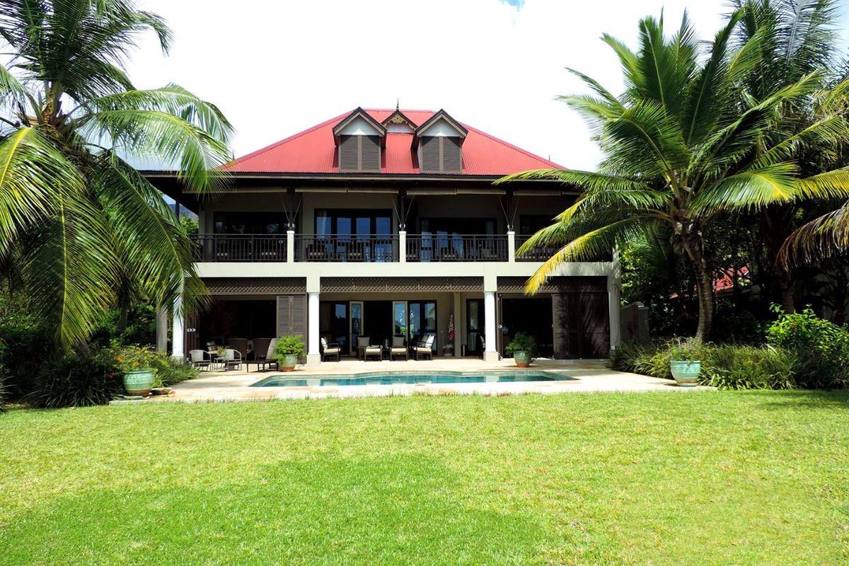 A two-story villa featuring a red metal roof, wrap-around balconies, and a ground-floor patio overlooking a private swimming pool and landscaped garden.