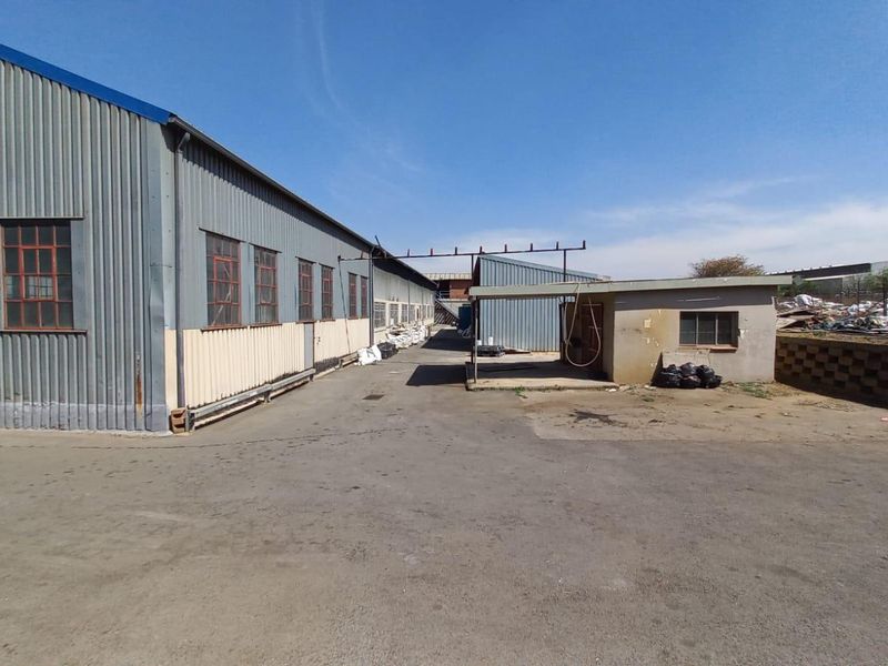 Industrial yard area featuring corrugated metal warehouse structures, paved asphalt surfacing, and a small covered utility or loading bay.