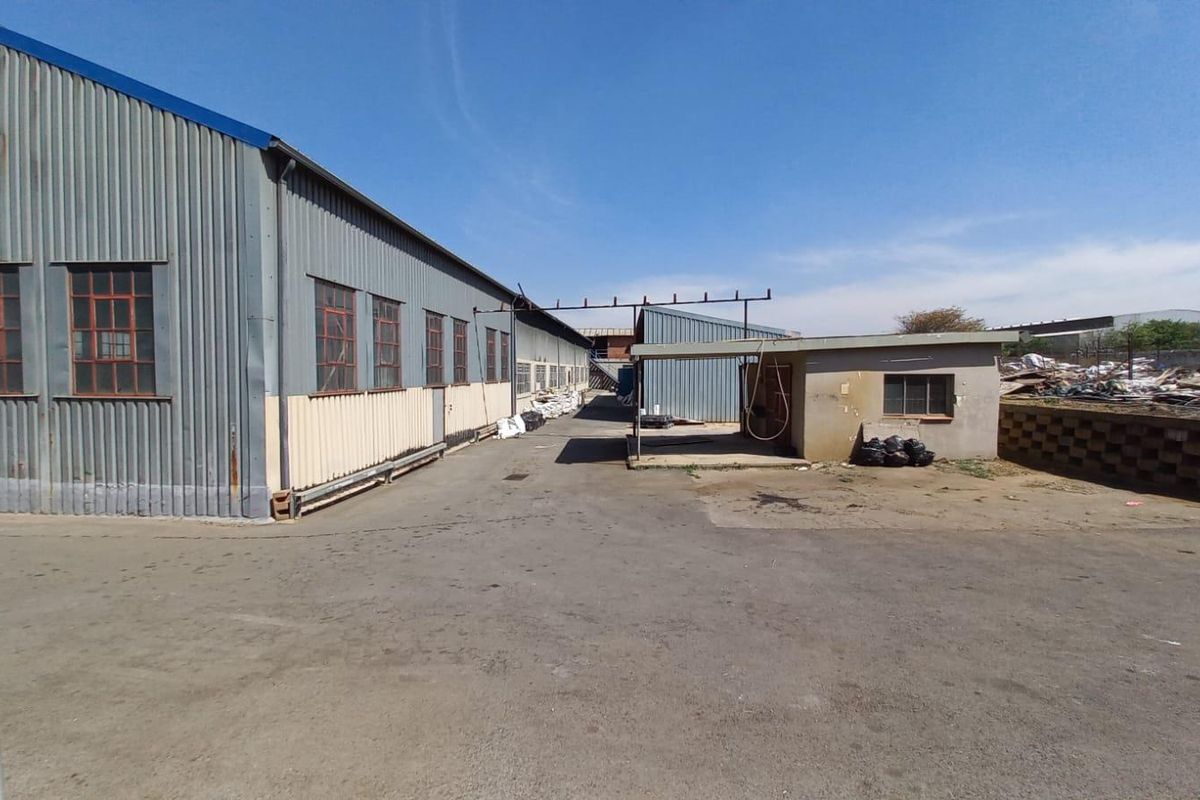 Industrial yard area featuring corrugated metal warehouse structures, paved asphalt surfacing, and a small covered utility or loading bay.