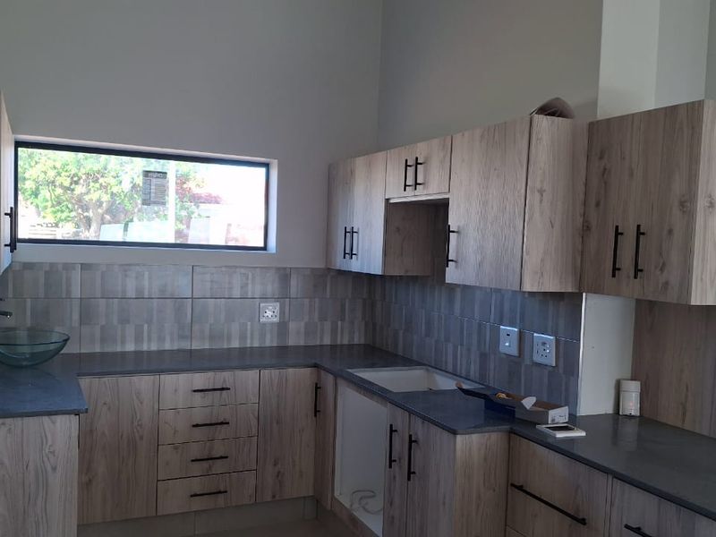 Modern kitchen featuring light wood-grain cabinetry, dark laminate countertops, tiled backsplash, and dual sink areas in a clean, empty space.
