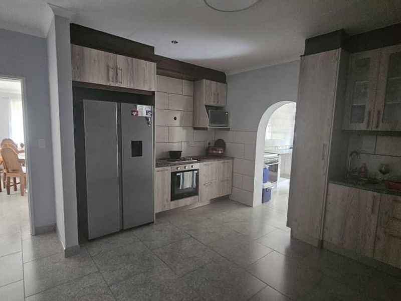 Modern kitchen featuring light wood-finish cabinetry, tiled backsplash, built-in oven and hob, and tiled flooring, with an adjacent archway leading to a dining area.