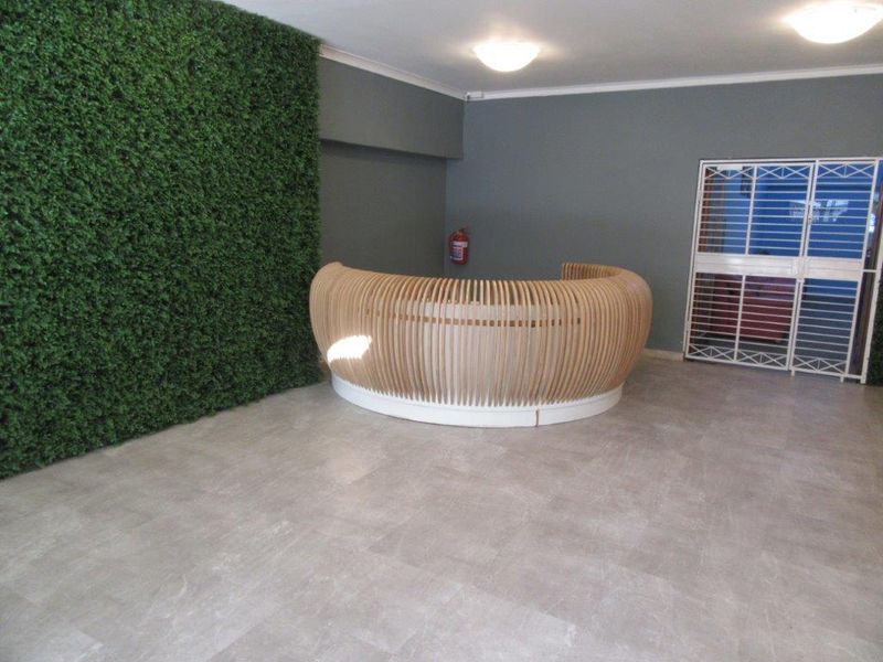 A modern entrance area featuring a curved wooden reception desk, a decorative artificial green wall, and tiled flooring.