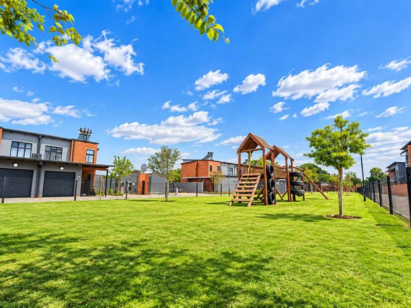 A well-maintained communal garden area featuring a manicured lawn, perimeter fencing, and a wooden children's play structure within a modern residential complex.