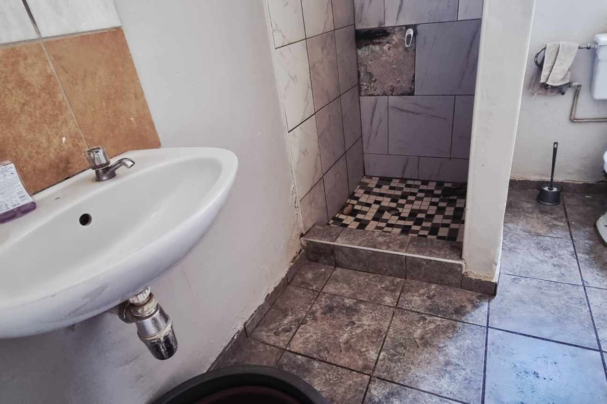 A functional bathroom featuring a wall-mounted basin, a shower cubicle with damaged tiling, and a standard toilet, requiring cosmetic updates and repairs.
