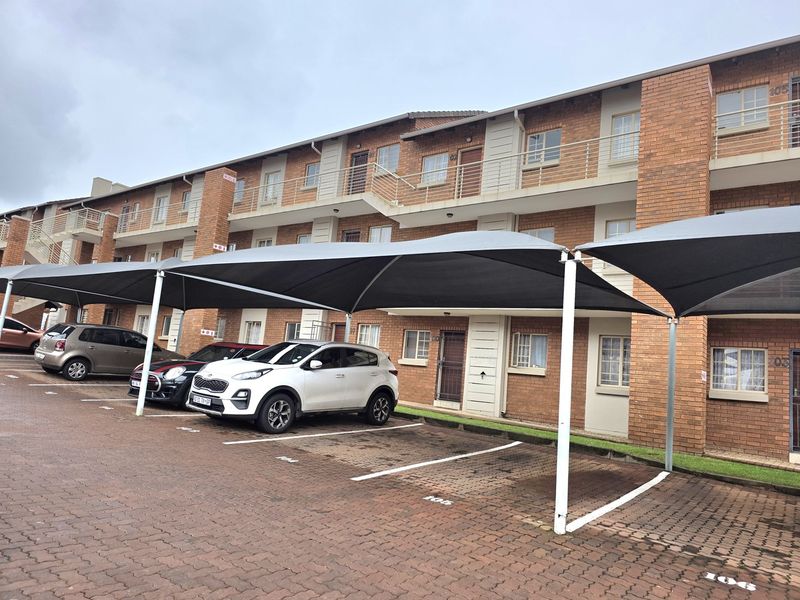 Exterior view of a multi-story apartment complex featuring brick facade, private balconies, and dedicated covered parking bays with shade cloth structures.