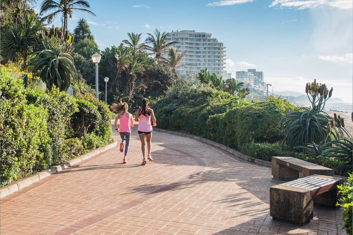 A scenic paved walkway lined with landscaped gardens and fixed seating, offering a pleasant outdoor environment near the coast.
