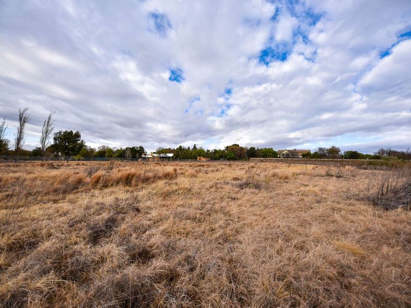 A wide-angle view of a vacant residential plot featuring dry grass and perimeter fencing under a cloudy sky.