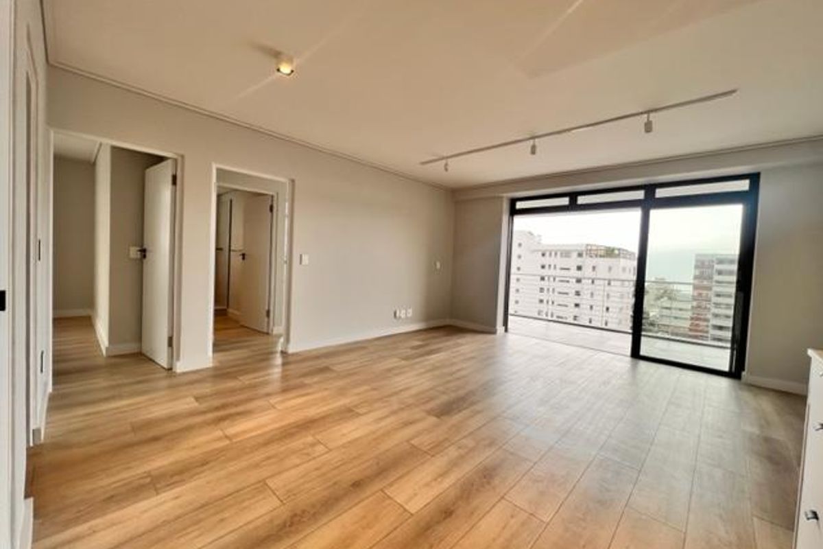 A bright, empty living room featuring modern laminate flooring, recessed and track lighting, and a large sliding glass door leading to a balcony.