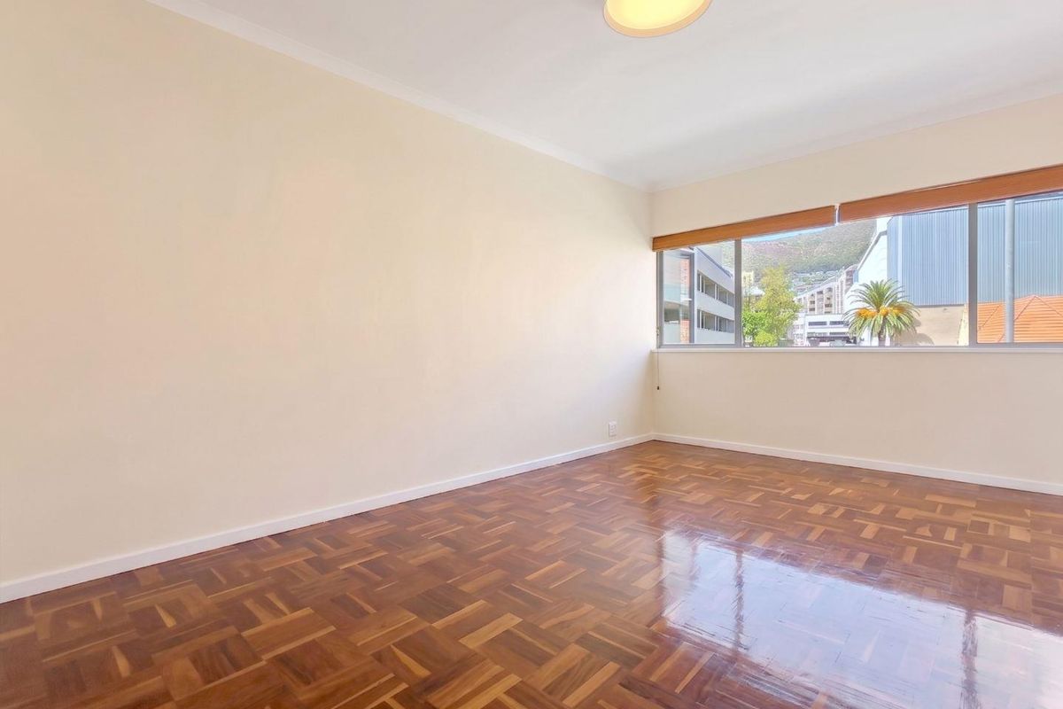 A bright, empty bedroom featuring polished parquet flooring, neutral walls, and a large window with fitted blinds.