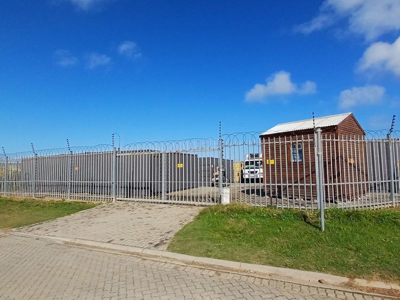 Industrial property exterior featuring secure perimeter fencing with razor wire, a paved entrance area, and a small timber utility structure.