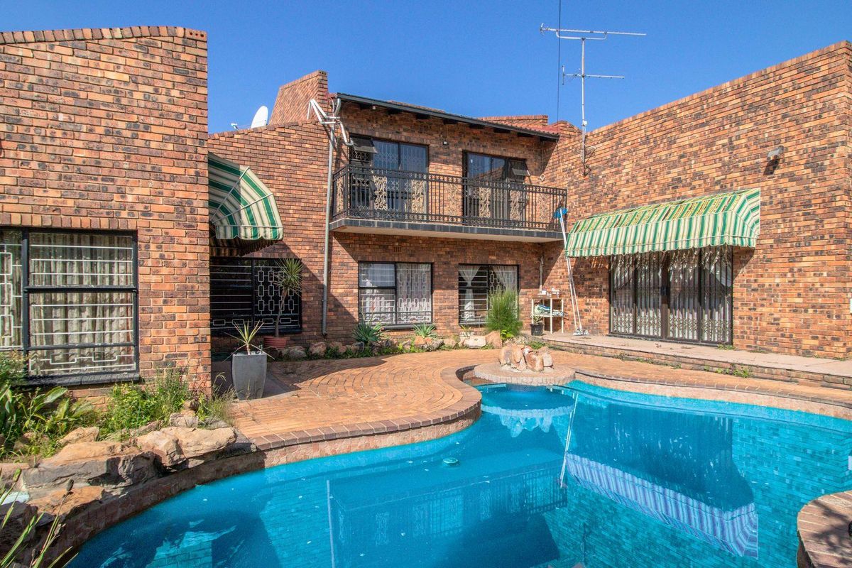 Rear exterior view of a double-story brick house featuring a swimming pool, paved patio area, and a second-floor balcony with metal railings.