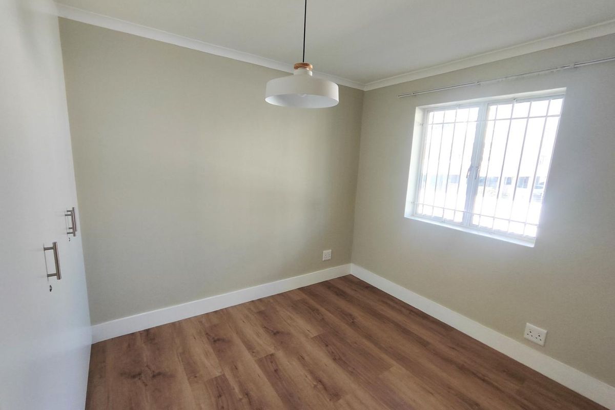 A bright, empty bedroom featuring modern wood-look laminate flooring, built-in cupboards, and a window fitted with security bars.
