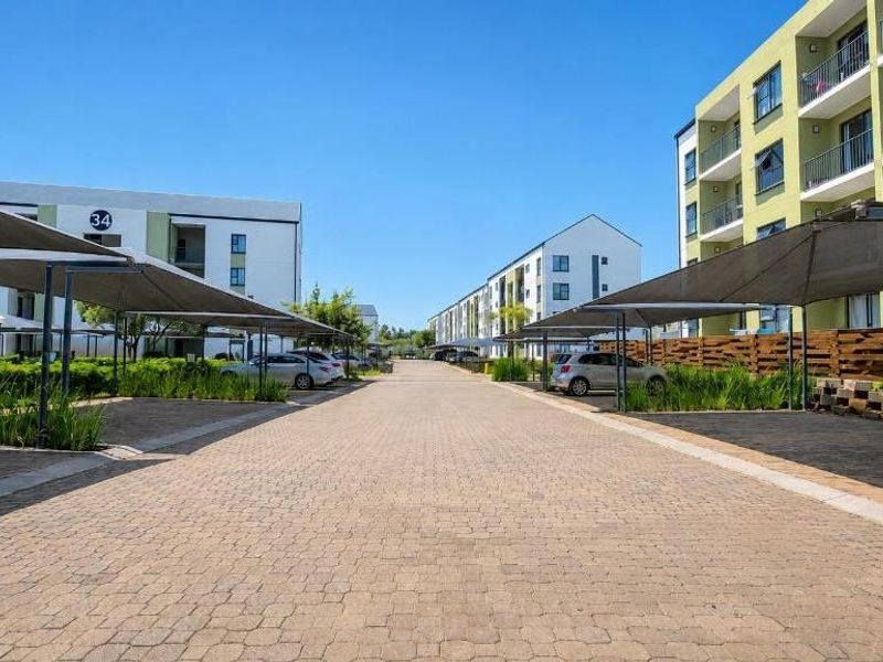 A well-maintained apartment complex featuring paved driveways, covered parking bays with shade cloth, and landscaped garden areas between the residential buildings.