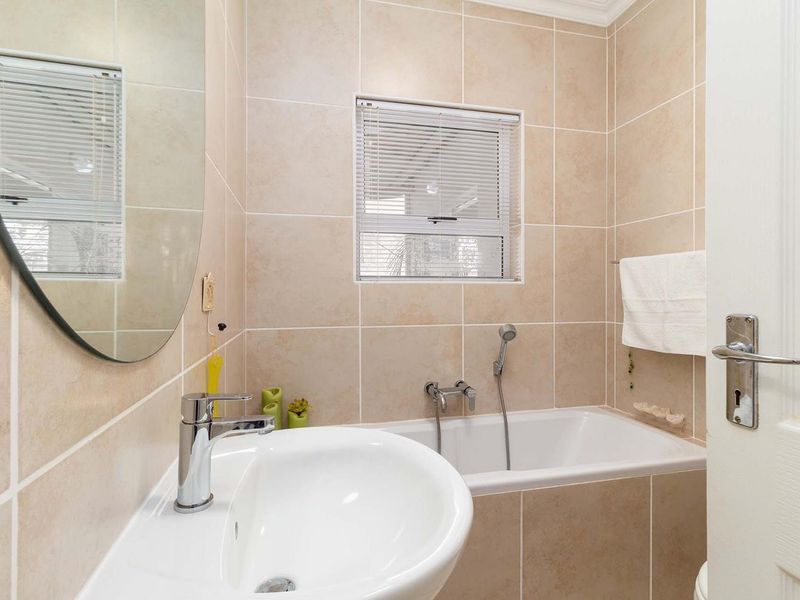 A compact, well-maintained bathroom featuring neutral ceramic wall tiles, a built-in bathtub with a handheld shower mixer, and a modern pedestal basin.