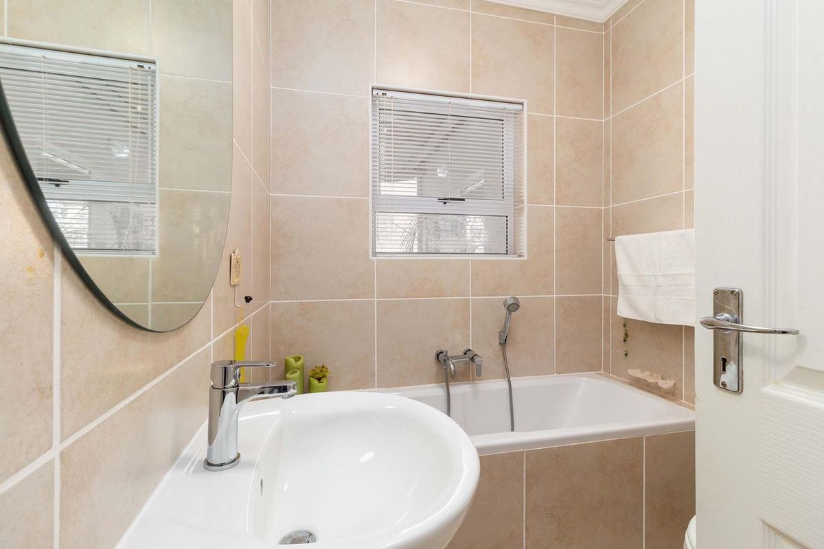 A compact, well-maintained bathroom featuring neutral ceramic wall tiles, a built-in bathtub with a handheld shower mixer, and a modern pedestal basin.