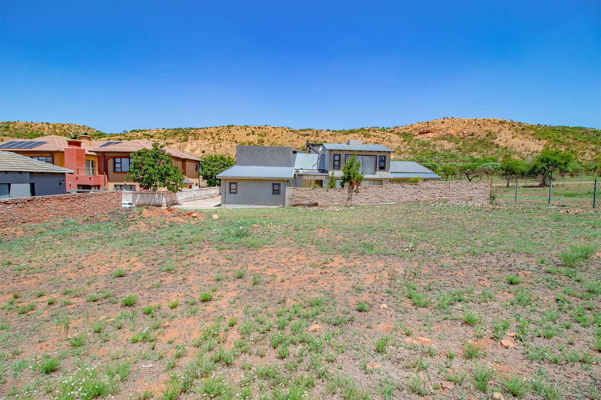 A view of a vacant residential plot with a boundary wall and neighboring properties in the background.