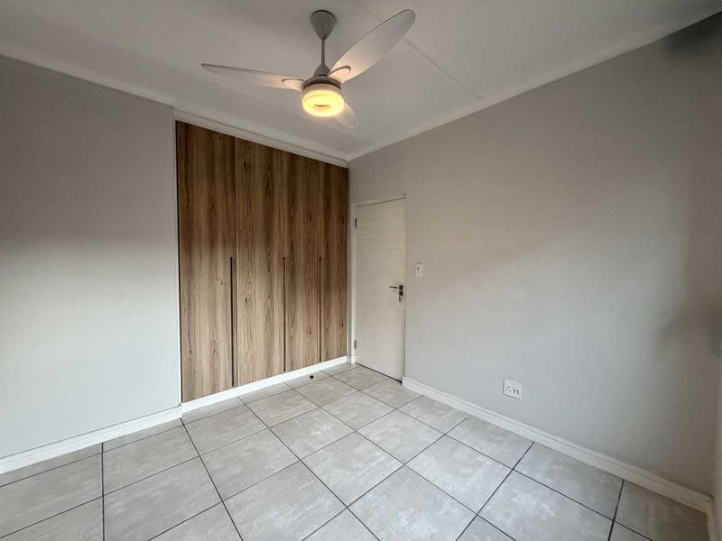 A clean, empty bedroom featuring modern wood-finish built-in cupboards, neutral tiled flooring, and a ceiling fan.