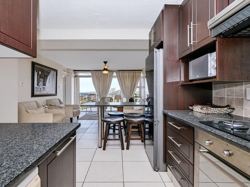 Open-plan kitchen and living area featuring dark wood cabinetry, granite countertops, built-in appliances, and tiled flooring, leading to a balcony with coastal views.
