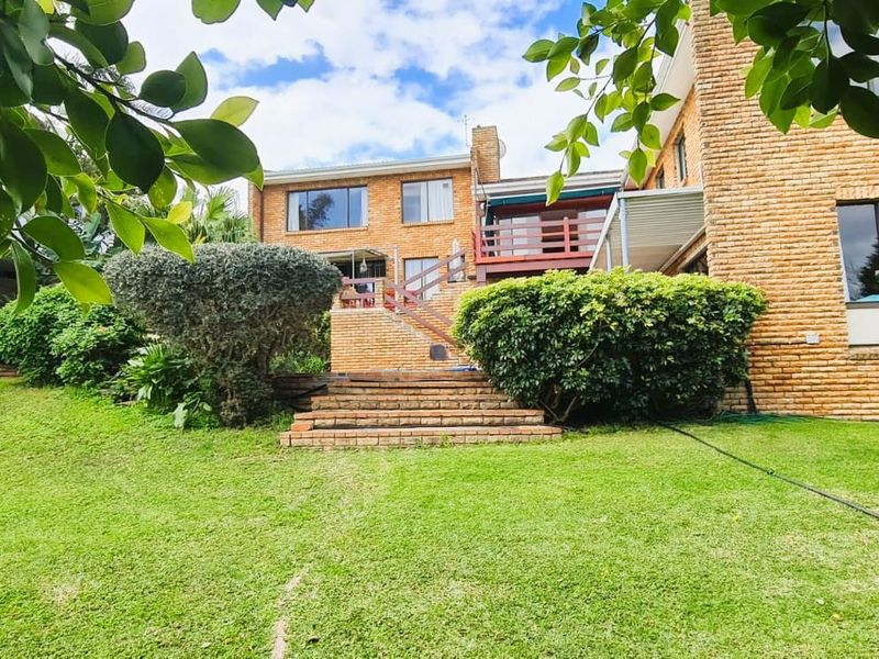 Rear view of the property featuring a spacious lawn, landscaped garden, brick facade, and a raised wooden deck accessible via a brick staircase.