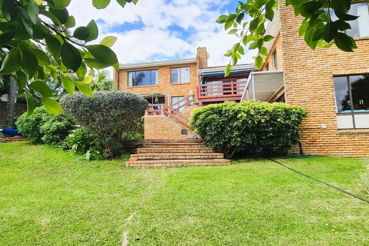 Rear view of the property featuring a spacious lawn, landscaped garden, brick facade, and a raised wooden deck accessible via a brick staircase.