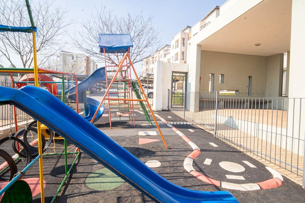 Communal outdoor play area featuring fixed playground equipment, rubberized flooring, and an adjacent covered patio space within the apartment complex.