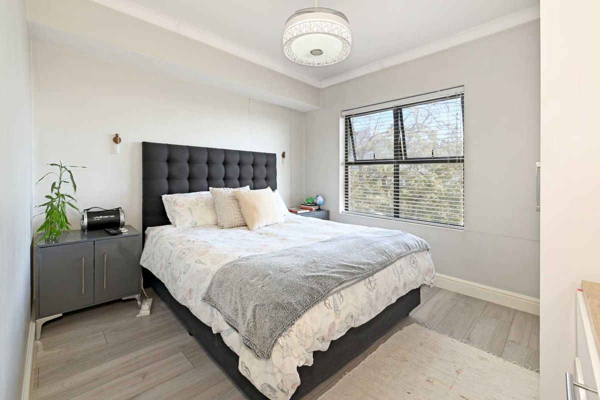 A well-lit bedroom featuring modern wood-look flooring, a large window with fitted blinds, and a contemporary ceiling light fixture.