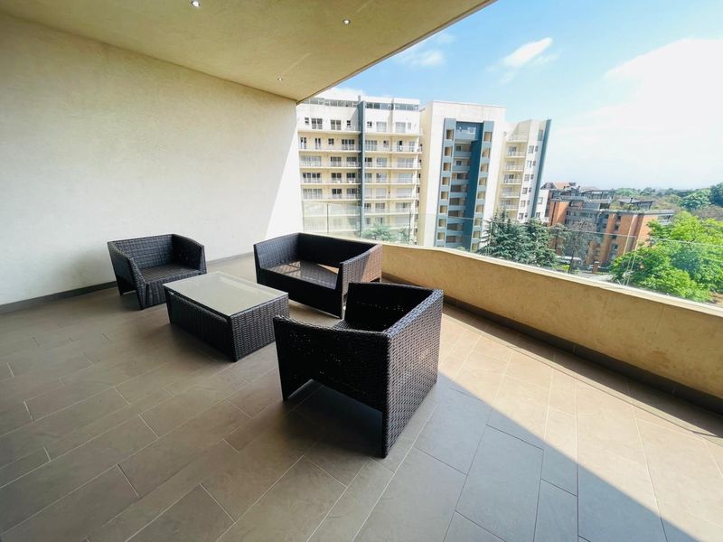 Spacious covered balcony featuring tiled flooring, recessed lighting, and a modern glass balustrade with views of the surrounding apartment complex.