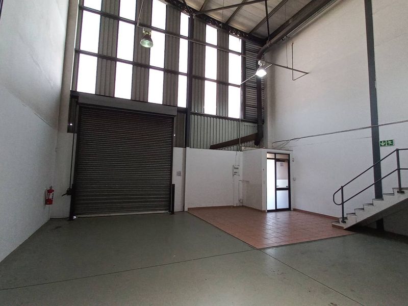 Spacious industrial warehouse unit featuring high ceilings, a large roller shutter door, mezzanine access stairs, and a dedicated tiled office or reception area.
