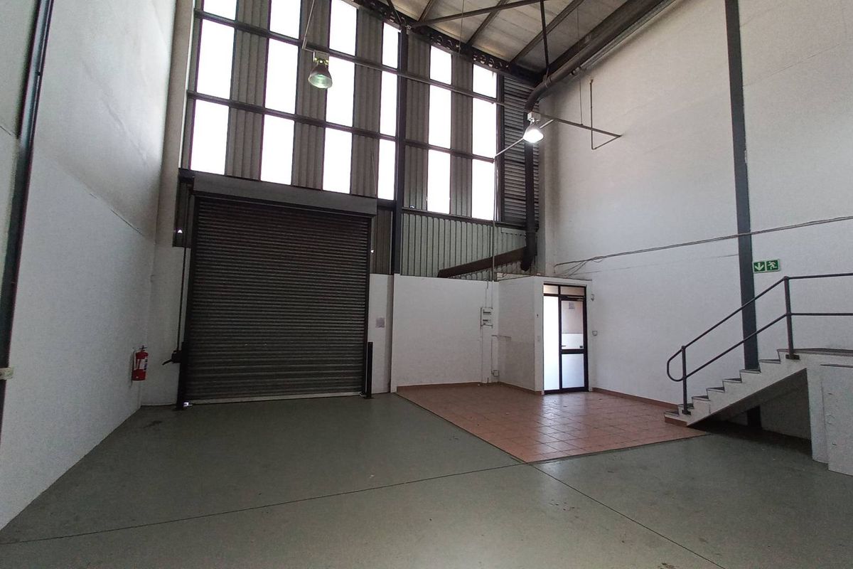 Spacious industrial warehouse unit featuring high ceilings, a large roller shutter door, mezzanine access stairs, and a dedicated tiled office or reception area.