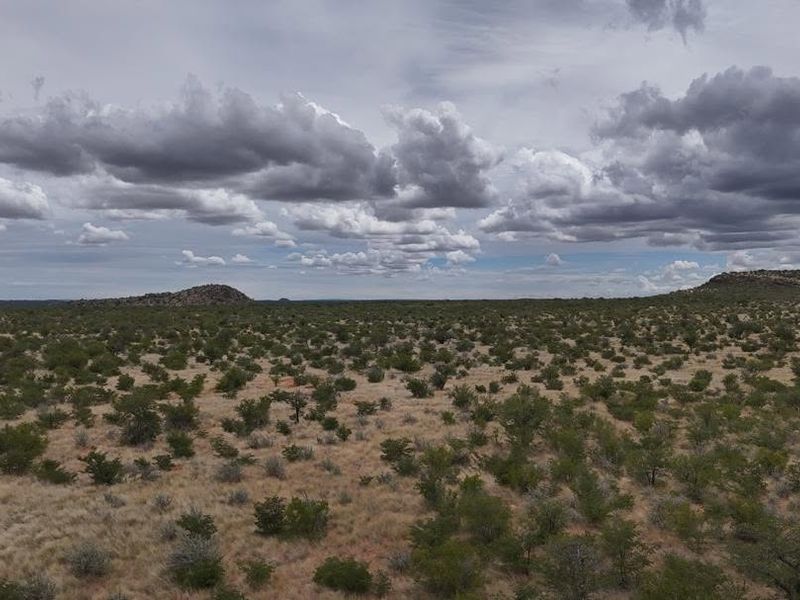 A wide-angle aerial view of the expansive farm landscape featuring natural vegetation, rolling terrain, and distant rocky outcrops under a cloudy sky.