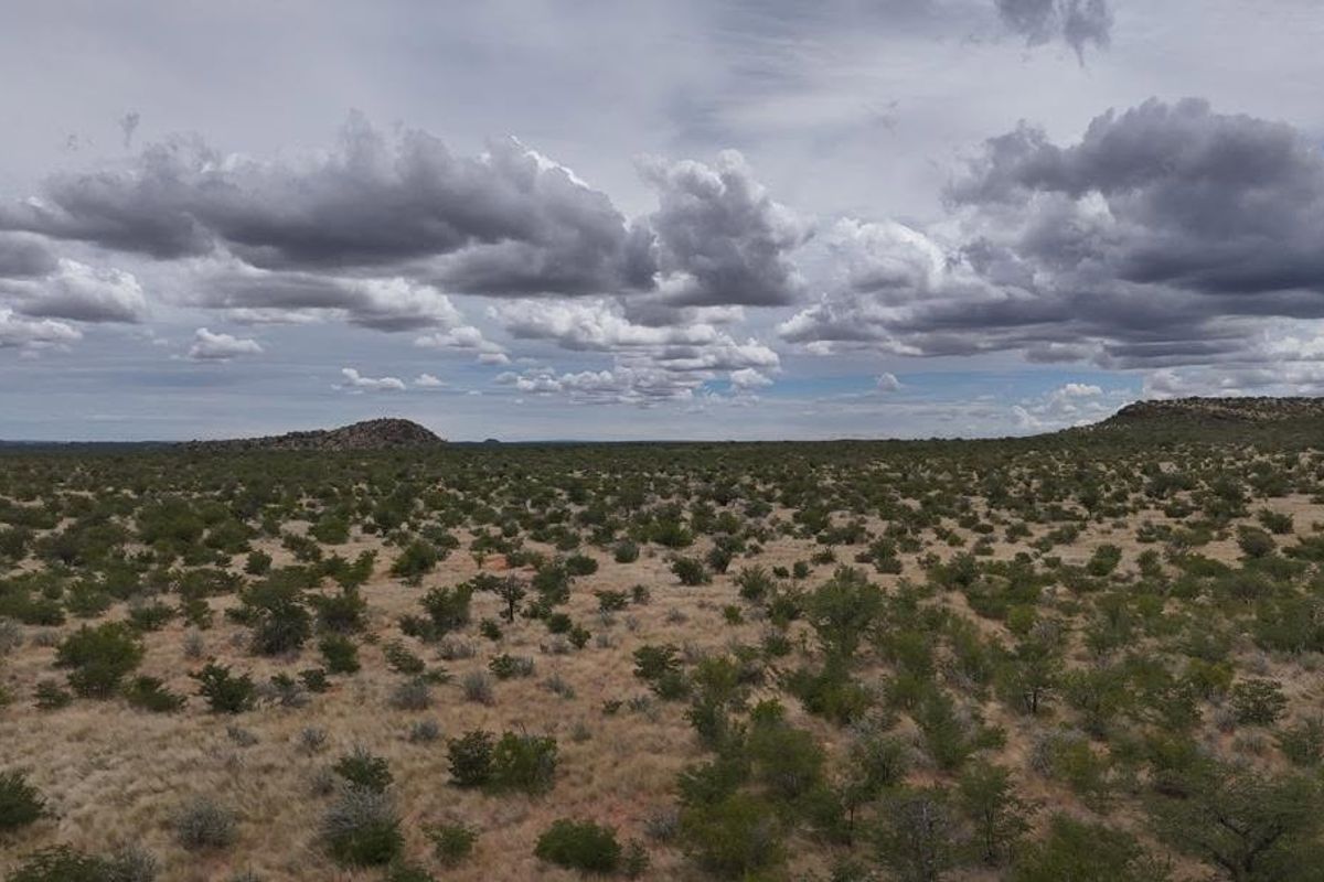 A wide-angle aerial view of the expansive farm landscape featuring natural vegetation, rolling terrain, and distant rocky outcrops under a cloudy sky.
