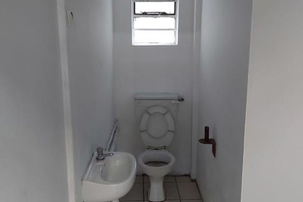 A compact industrial restroom featuring a wall-mounted basin, standard toilet, and tiled flooring with a small window for ventilation.