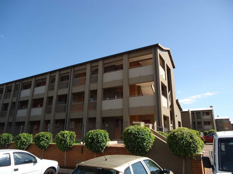 Exterior view of a multi-story apartment complex featuring a modern facade, balcony levels, and a brick perimeter wall with landscaped shrubs.
