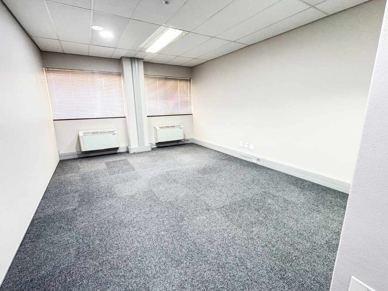A clean, empty office space featuring grey carpet flooring, a suspended ceiling with recessed lighting, and two wall-mounted HVAC units.