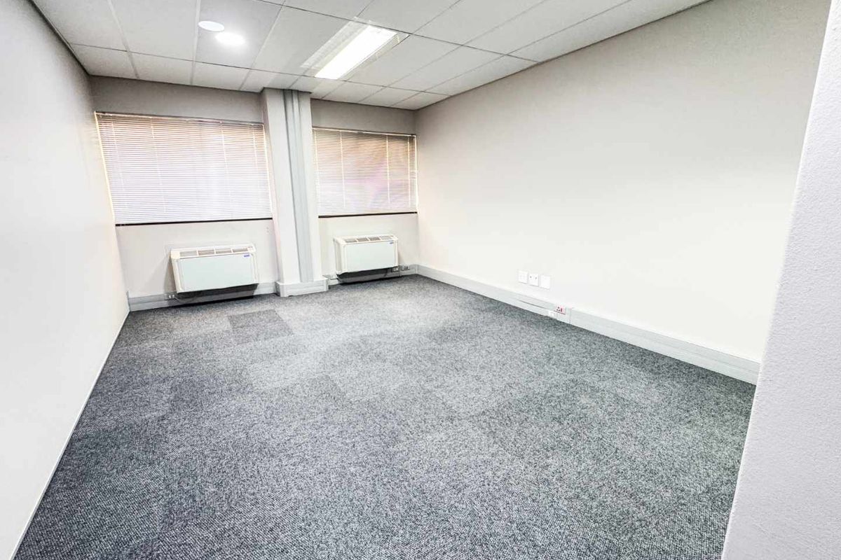 A clean, empty office space featuring grey carpet flooring, a suspended ceiling with recessed lighting, and two wall-mounted HVAC units.