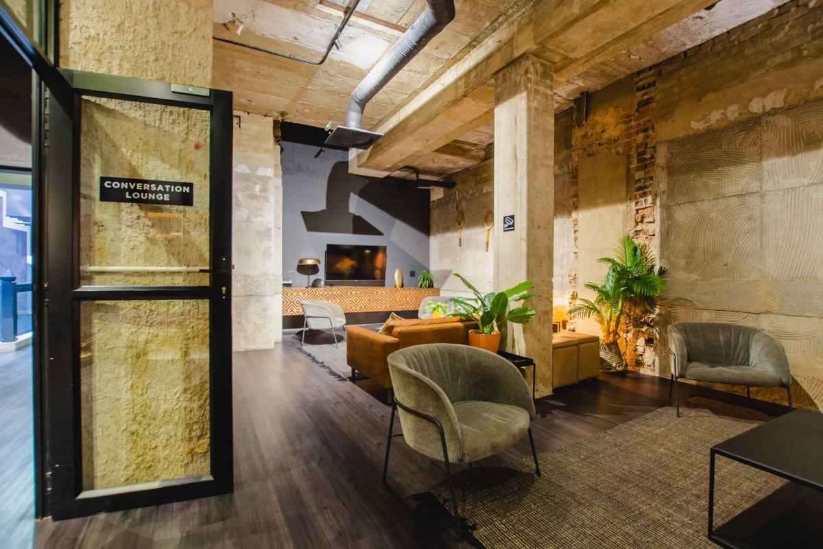 A modern industrial-style conversation lounge featuring exposed concrete ceilings, raw textured walls, and dark wood-look flooring in a well-maintained communal apartment space.