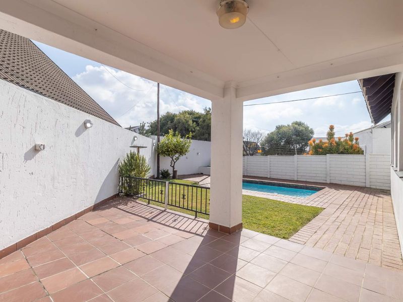 Covered patio area overlooking a private garden and swimming pool, featuring tiled flooring and a structural support pillar.