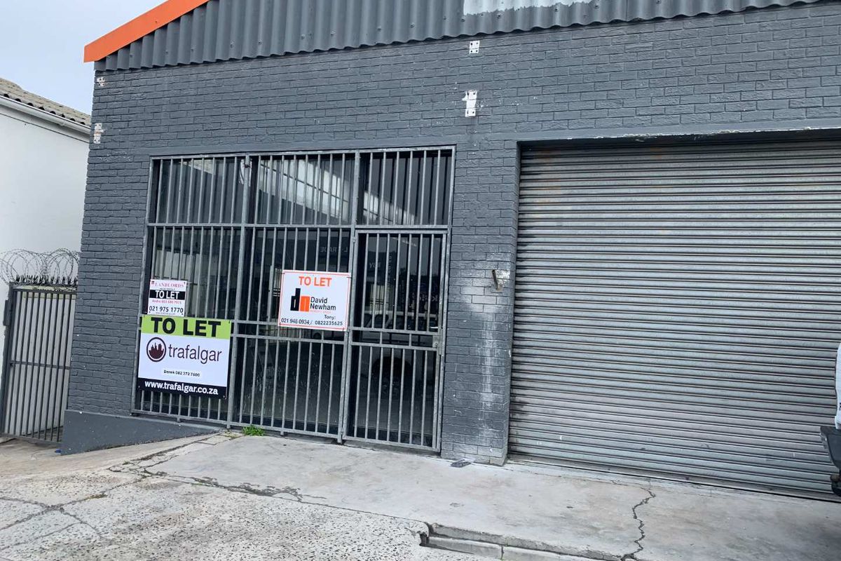Front facade of a retail or industrial unit featuring a roller shutter door, security gate, and brick exterior with visible signage.