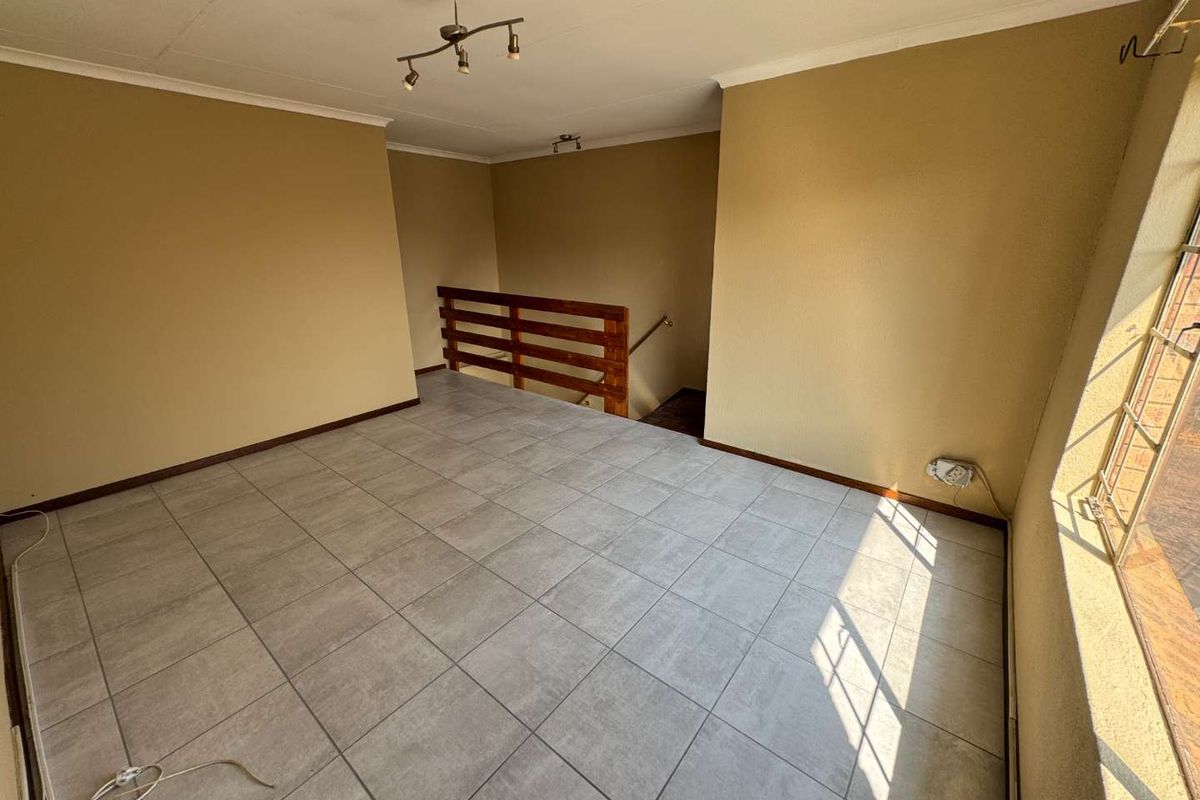 An empty, tiled living area featuring a wooden staircase railing, neutral wall tones, and natural light from a window with security bars.