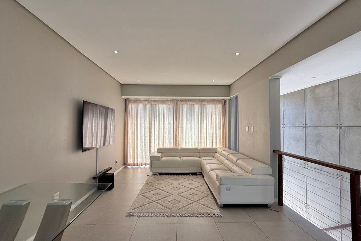 Modern living area featuring tiled flooring, recessed ceiling lighting, and a feature wall with concrete-style paneling and a staircase balustrade.